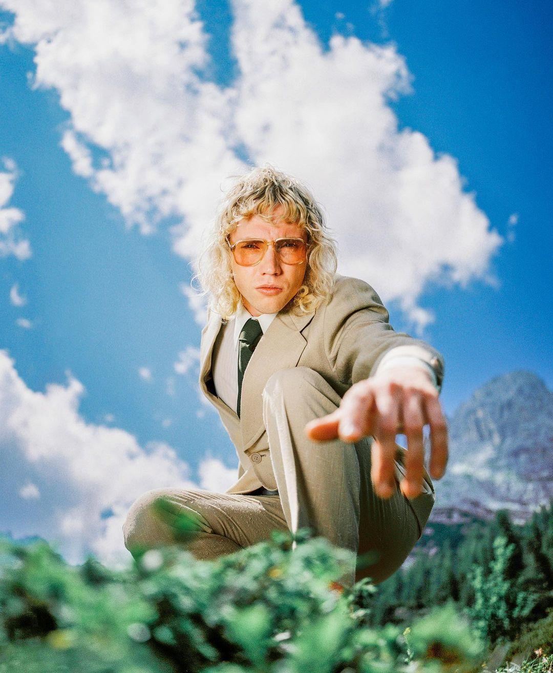 Man in a tan suit against a saturated blue sky, reaching toward camera