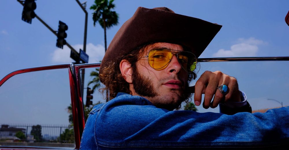 Andrea Calvetti in a cowboy hat and yellow sunglasses, leaning out of a vintage convertible under LA palm trees